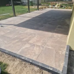 Wykończona powierzchnia z płyt tarasowych w odcieniach beżu, obramowana ciemnoszarą kostką. Widoczny trawnik, elementy konstrukcji zadaszenia i budynek w tle.