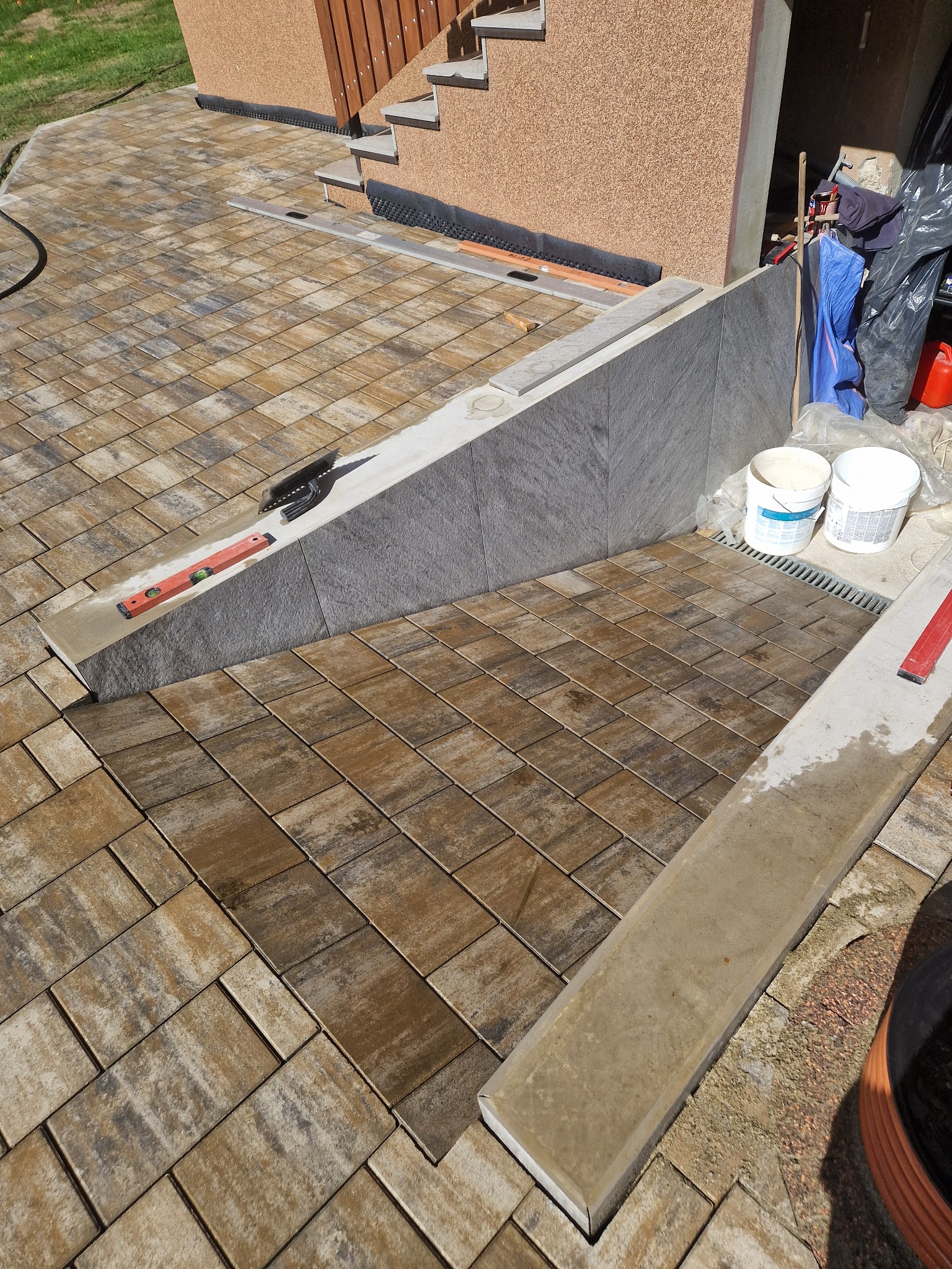 Ułożona kostka brukowa w odcieniach brązu i beżu, widoczne narzędzia: poziomica i kielnia, oraz fragment budynku z schodami i elementami odwodnienia liniowego.