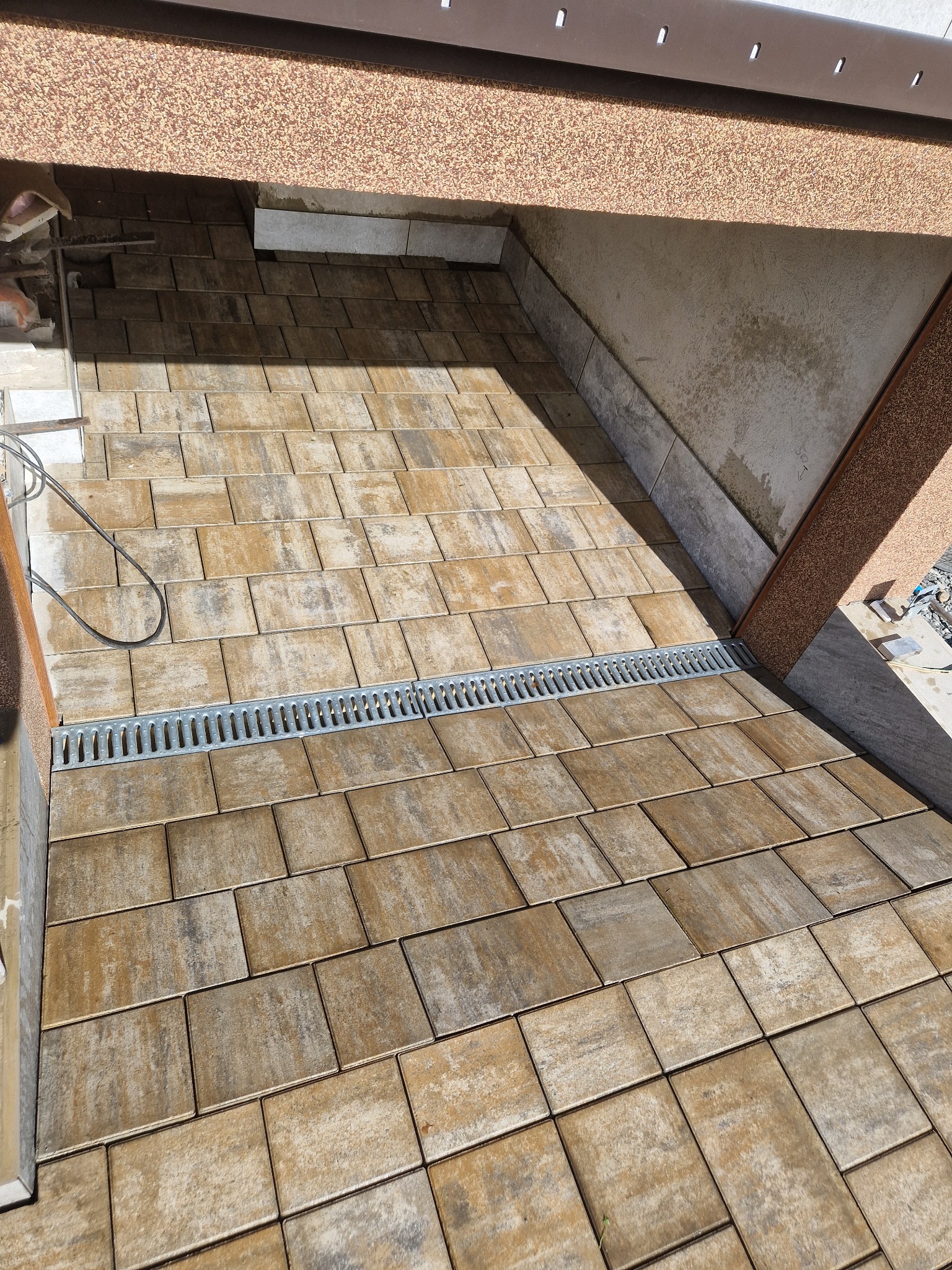 Ułożona kostka brukowa w odcieniach beżu i brązu z odpływem liniowym, widok z góry pod zadaszeniem.