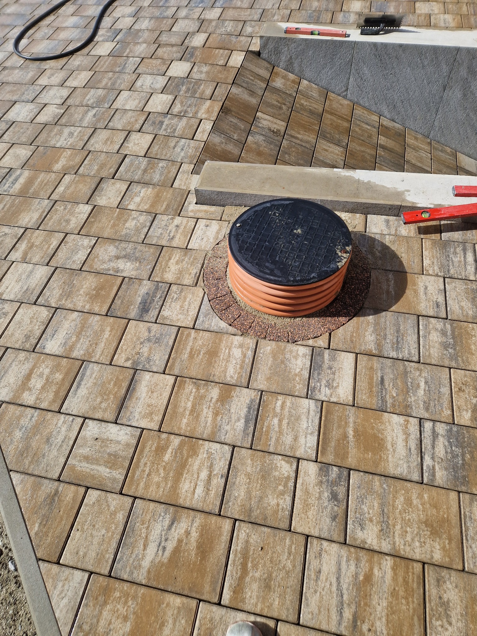 Ułożona kostka brukowa w odcieniach brązu i szarości, z widocznym włazem kanalizacyjnym otoczonym drobnym żwirem, poziomice oraz czarny przewód w tle.