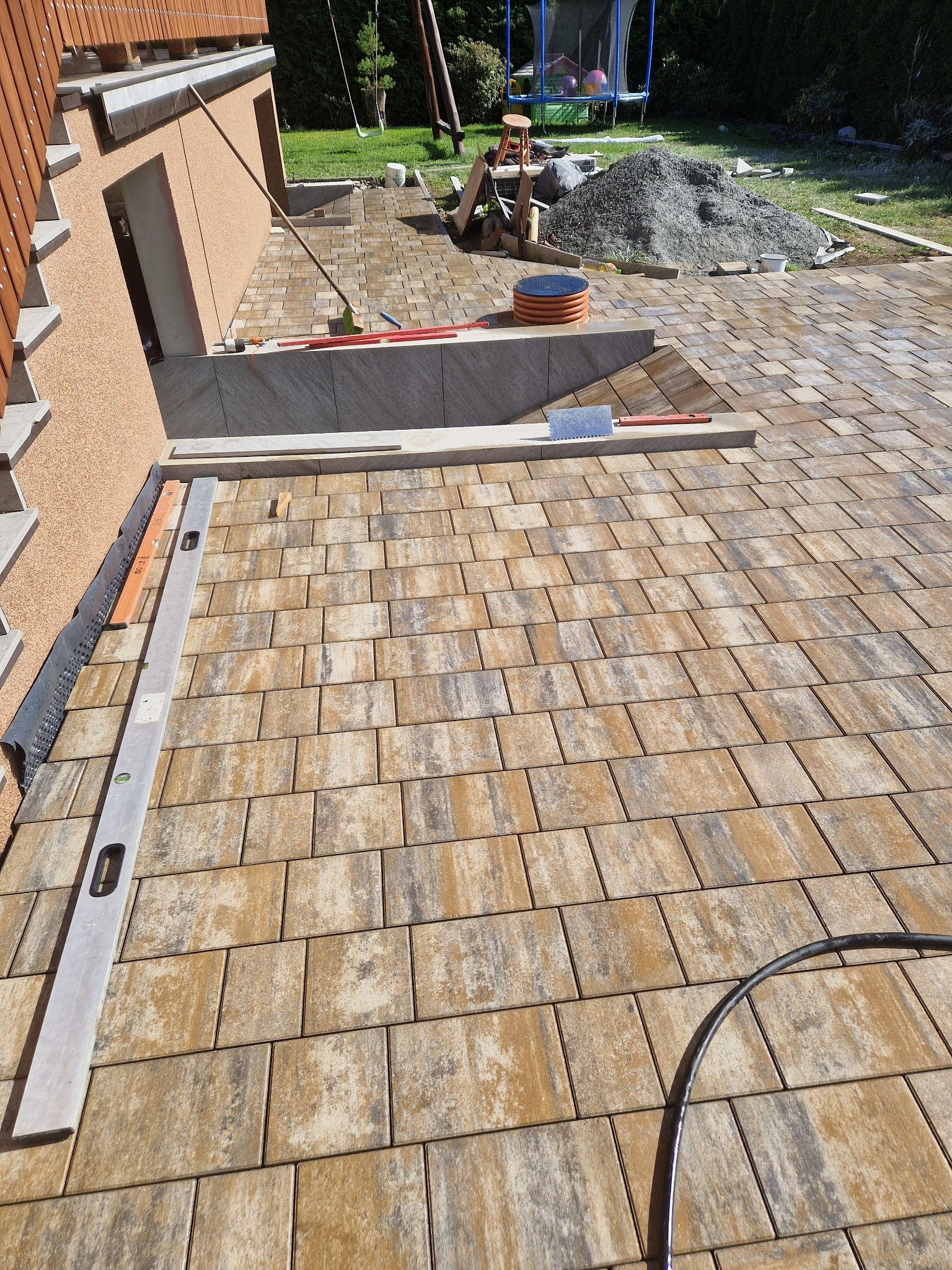 Ułożona częściowo kostka brukowa w odcieniach beżu i szarości, widoczny poziomica, łata oraz stos materiału sypkiego w tle, na trawniku