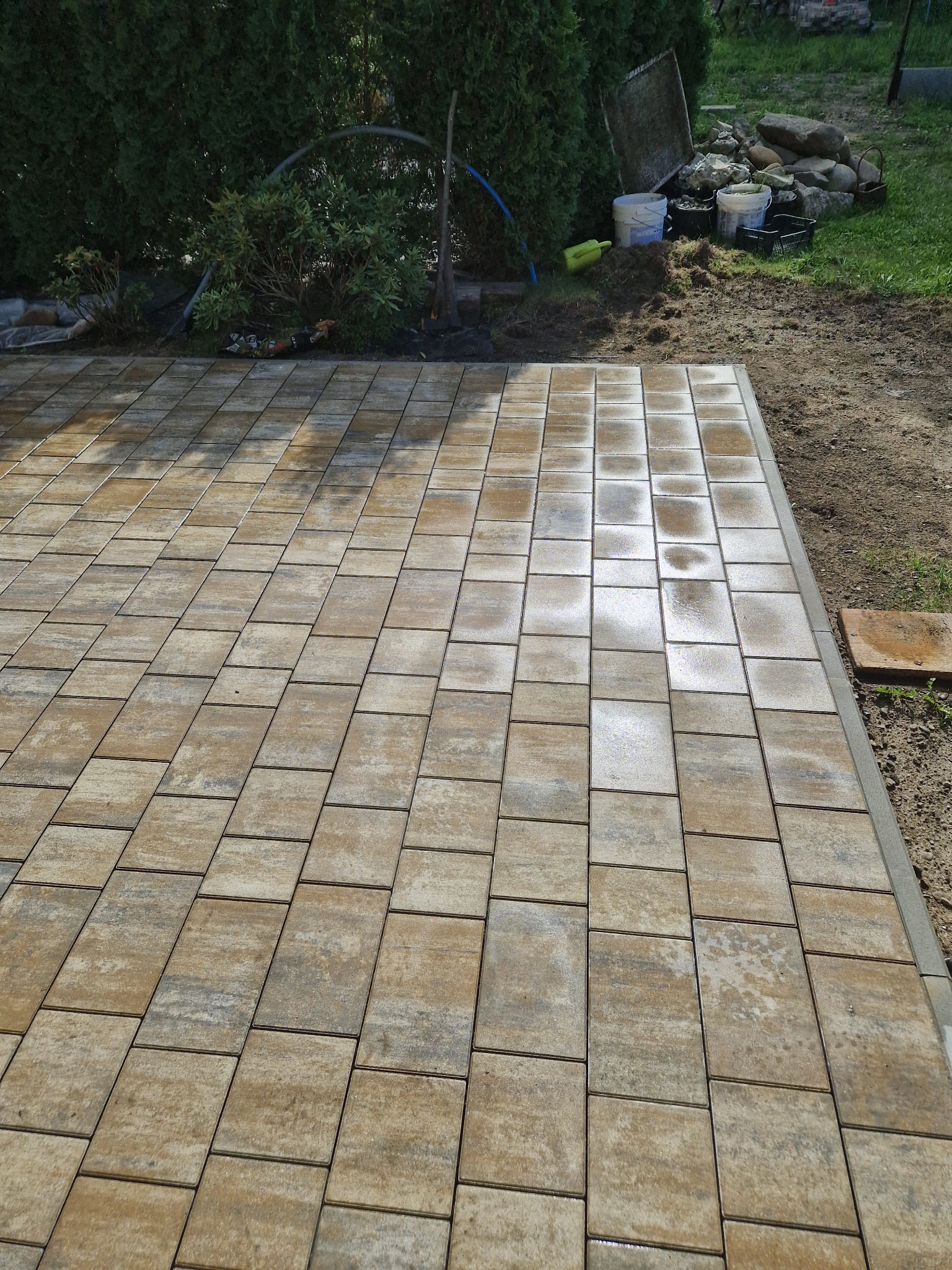 Świeżo ułożona, brązowo-szara kostka brukowa na posesji, z widocznymi śladami wilgoci i niedokończonym obrzeżem z prawej strony, tło z zielenią i narzędziami.