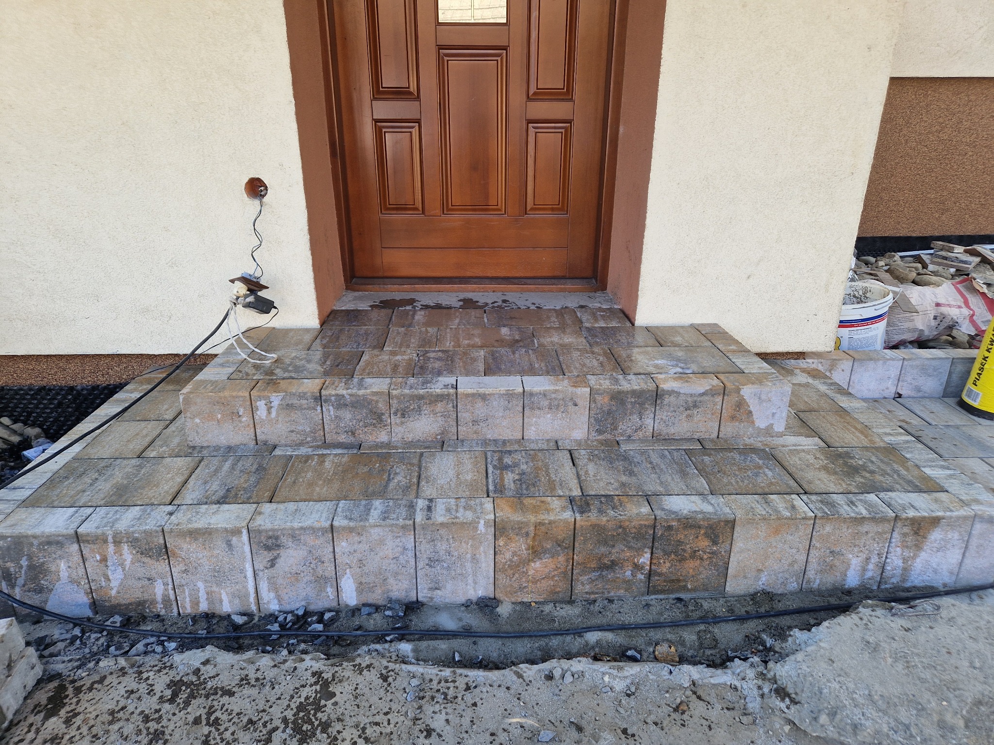 Nowe, trójstopniowe schody wejściowe z kostki brukowej w odcieniach szarości i brązu, prowadzące do drewnianych drzwi wejściowych. Widoczne niedokończone prace wykończeniowe wokół schodów...