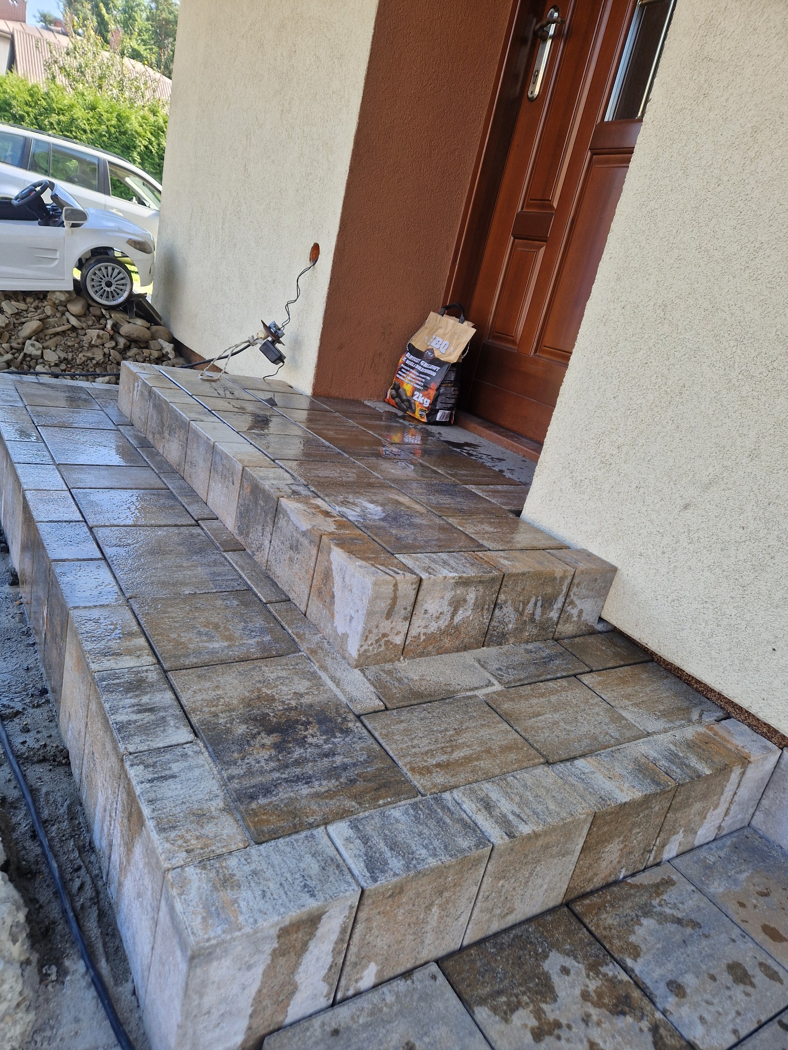 Ułożone schody wejściowe z kostki brukowej w Pruchna, prowadzące do brązowych drzwi, widoczny worek z węglem do grilla i zabawkowy samochód.
