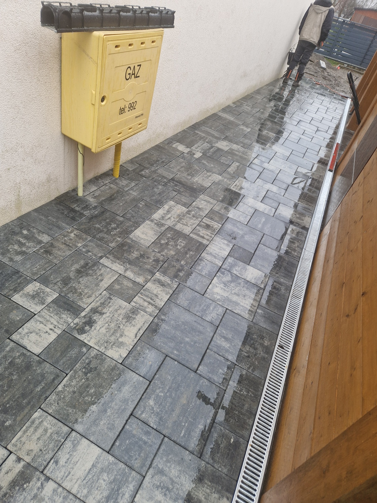 Ułożona, mokra kostka brukowa w odcieniach szarości i beżu, przylegająca do ściany z żółtą skrzynką gazową i odpływem liniowym wzdłuż drewnianego ogrodzenia. W tle widoczna postać w roboczym ubraniu.