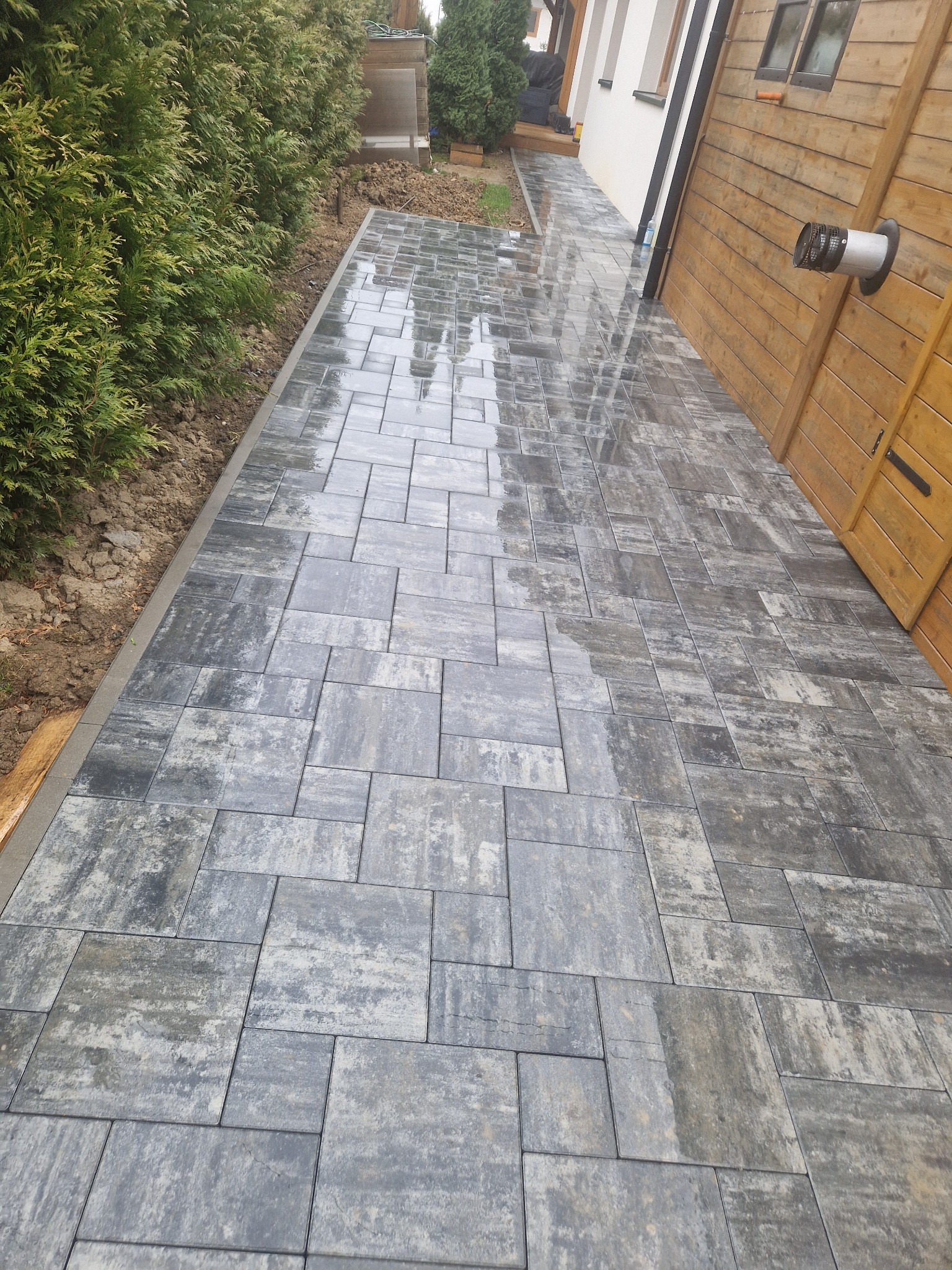 Świeżo ułożony chodnik z szarej kostki brukowej o nieregularnym wzorze, delikatnie mokry, przylegający do tujowego żywopłotu i drewnianej ściany budynku z widocznym kominem wentylacyjnym.