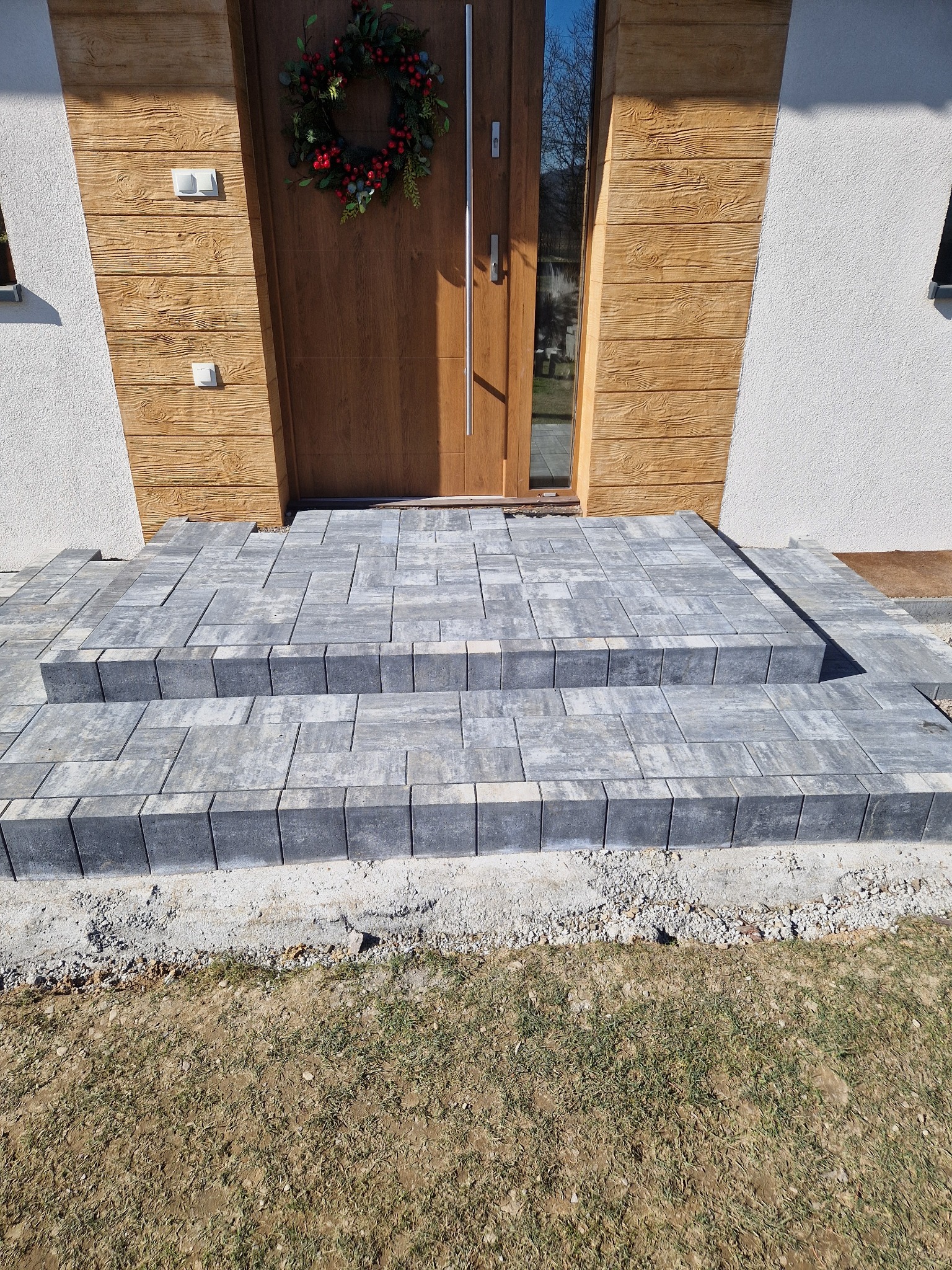 Schody wejściowe ułożone z szarej kostki brukowej przed drewnianymi drzwiami z wieńcem świątecznym, widoczne niedokończone obrzeże i trawnik.