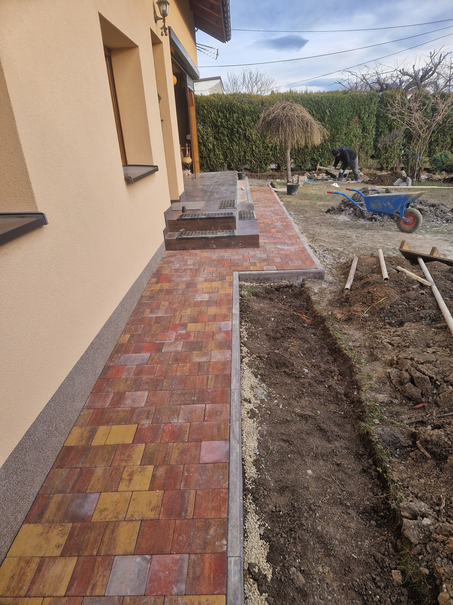 Ścieżka z wielobarwnej kostki prowadząca od domu, z widocznym wykopem pod kolejne prace brukarskie i pracownikiem w tle.