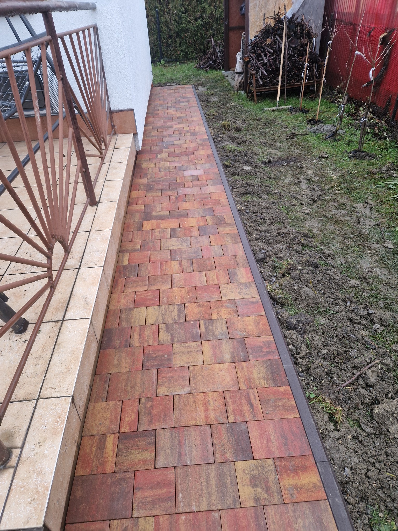 Świeżo ułożona ścieżka z kostki w odcieniach czerwieni i brązu, prowadząca wzdłuż ściany budynku z brązową barierką, obok rabata z ziemią i młodymi drzewkami.