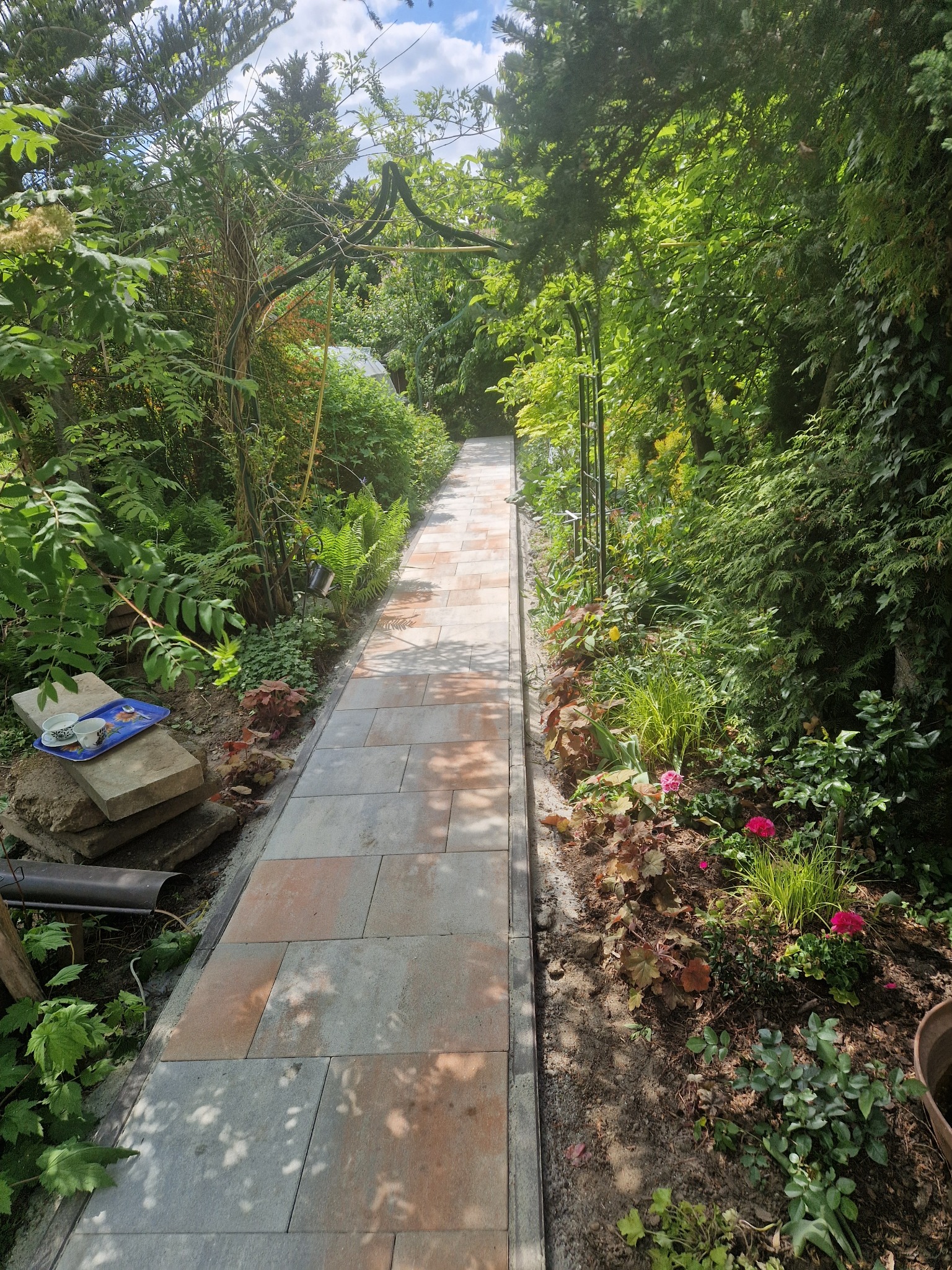 Ułożona ścieżka z kostki brukowej w ogrodzie, otoczona zielenią. Na pierwszym planie widoczny fragment tacy z filiżankami i elementami dekoracyjnymi.
