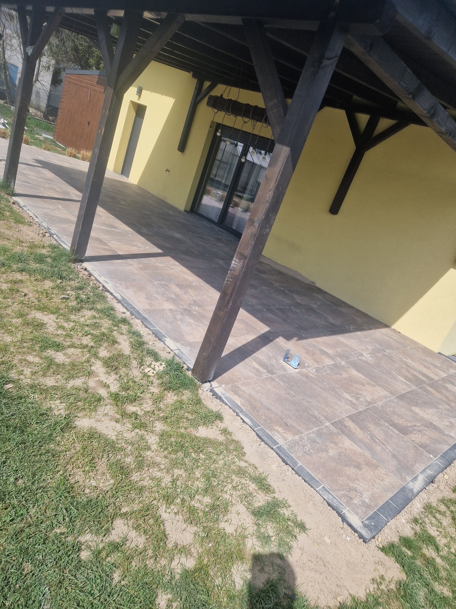 Wykończony taras z brązowych płyt, obramowany ciemną kostką, przylegający do żółtej ściany budynku z drewnianą konstrukcją zadaszenia. Widoczna trawa i fragment ogrodu.