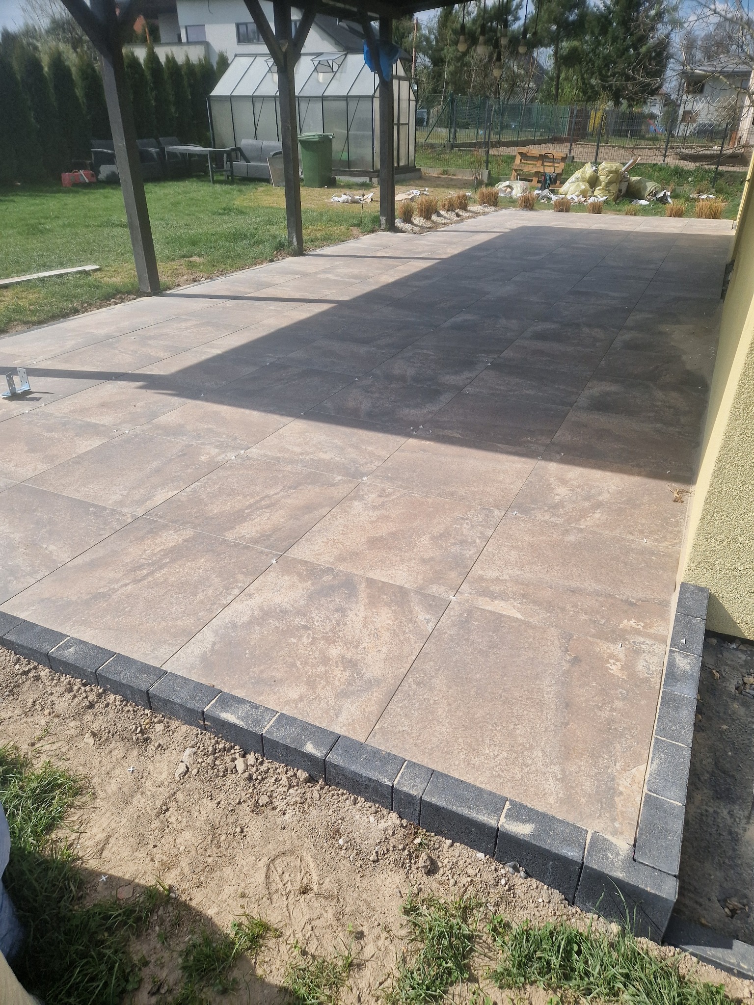 Wykończona powierzchnia z płyt tarasowych w odcieniach beżu, obramowana ciemnoszarą kostką. Widoczny trawnik, elementy konstrukcji zadaszenia i budynek w tle.