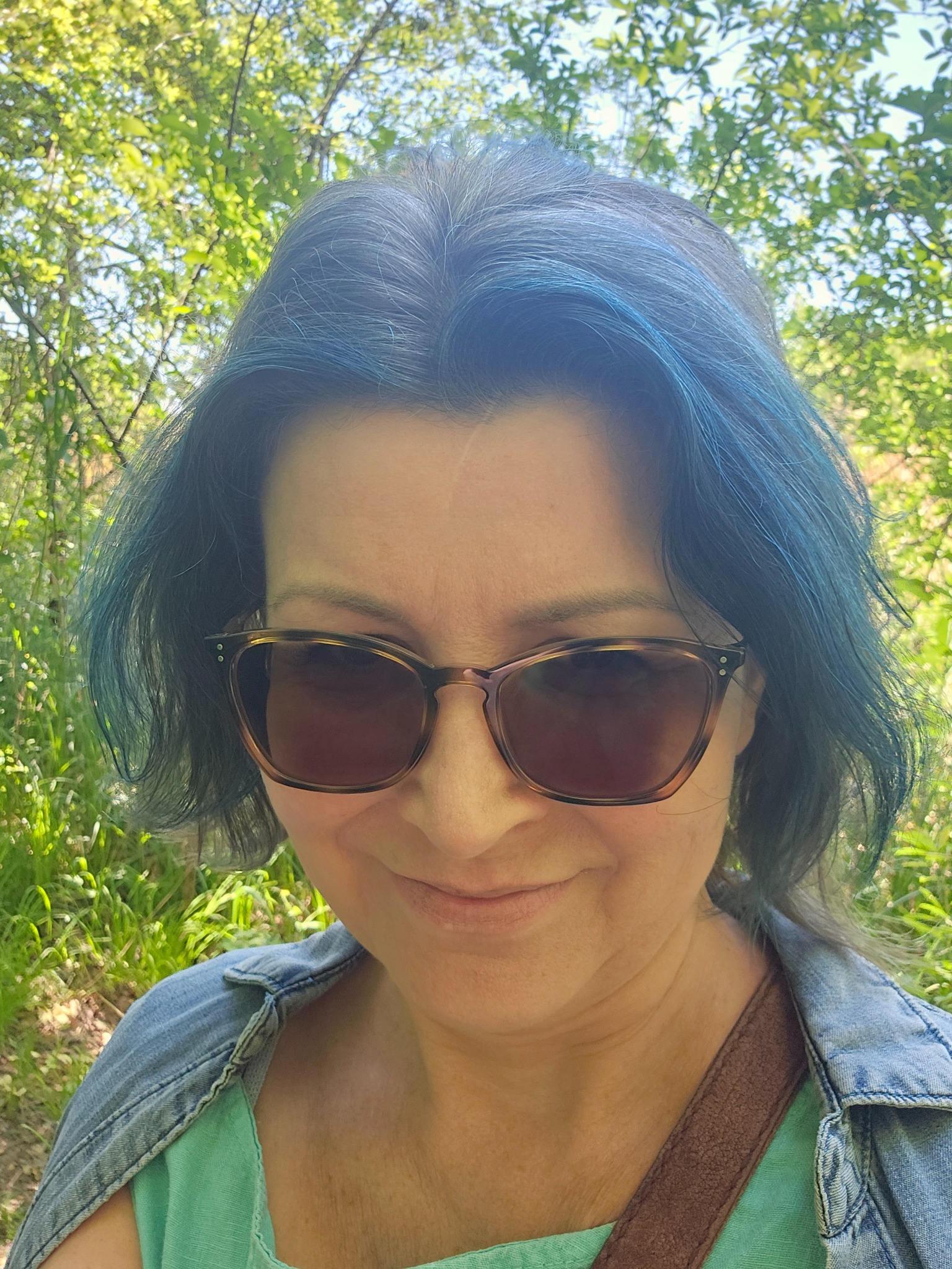 Portret kobiety w okularach przeciwsłonecznych, z niebieskimi pasemkami we włosach, na tle zieleni. Uśmiech, swobodny strój.