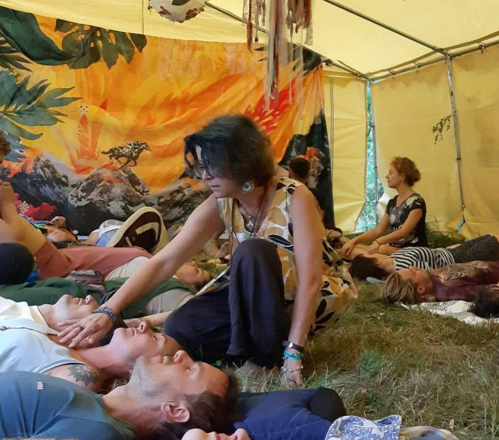 Grupowa sesja relaksacyjna w namiocie: osoby leżą na trawie, kobieta dotyka delikatnie klatki piersiowej jednej z nich. Na ścianie namiotu kolorowy obraz.