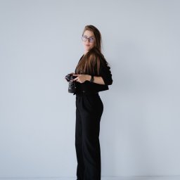 Noglik Photo - Kobieta z długimi włosami i okularami w czarnym stroju trzyma aparat fotograficzny, stojąc na tle białej ściany.