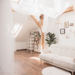 Noglik Photo - Jasne, przestronne poddasze z drewnianą podłogą, sofą w stylu vintage, drabiną z kwiatami i dekoracyjnymi lampami wiszącymi pod oknami dachowymi.