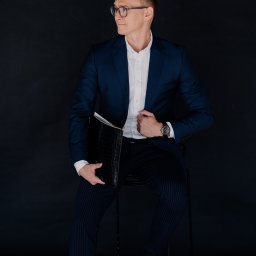 Noglik Photo - Mężczyzna w okularach, granatowym garniturze i białej koszuli, siedzący na krześle z czarną aktówką, spoglądający w bok.