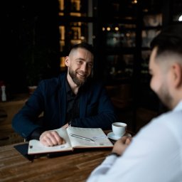 Noglik Photo - Mężczyzna w ciemnej marynarce uśmiecha się podczas spotkania biznesowego w kawiarni, notując coś w otwartym kalendarzu obok filiżanki kawy i długopisu.