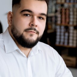 Noglik Photo - Portret brodatego mężczyzny w białej koszuli, z delikatnym uśmiechem, na rozmytym tle regałów.