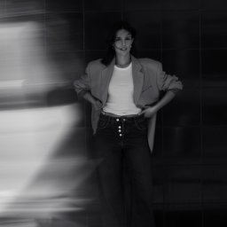 Noglik Photo - Portret kobiety w luźnym blazerze i jeansach z guzikami, pozującej na tle ciemnej, kafelkowanej ściany z efektem rozmycia po lewej stronie.