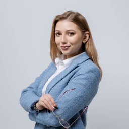 Noglik Photo - Uśmiechnięta blondynka w błękitnym tweedowym żakiecie i białej koszuli, trzymająca okulary w ręce, pozuje na jasnoszarym tle.