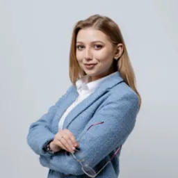 Uśmiechnięta blondynka w błękitnym tweedowym żakiecie i białej koszuli, trzymająca okulary w ręce, pozuje na jasnoszarym tle.