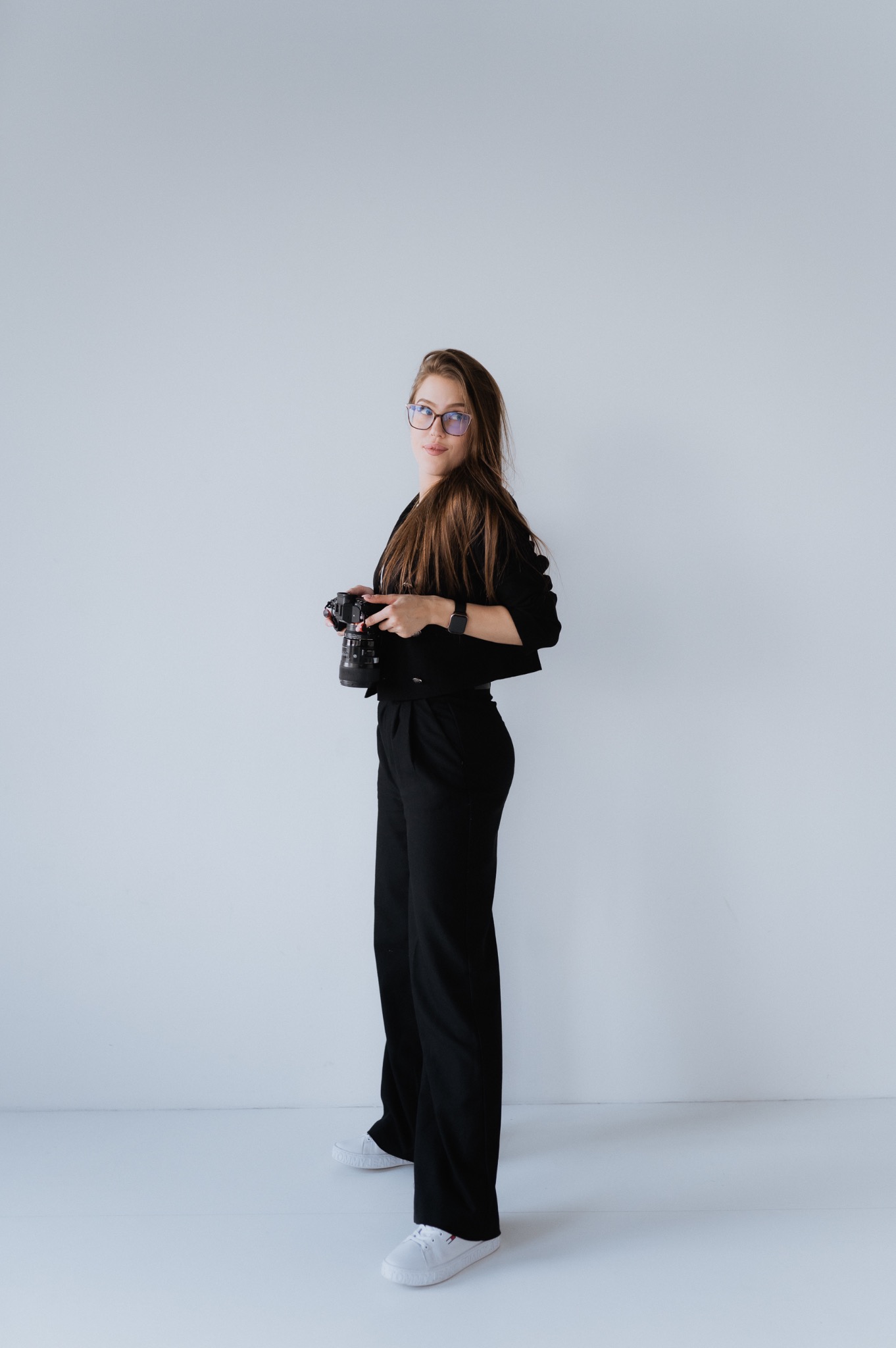 Kobieta z długimi włosami i okularami w czarnym stroju trzyma aparat fotograficzny, stojąc na tle białej ściany.