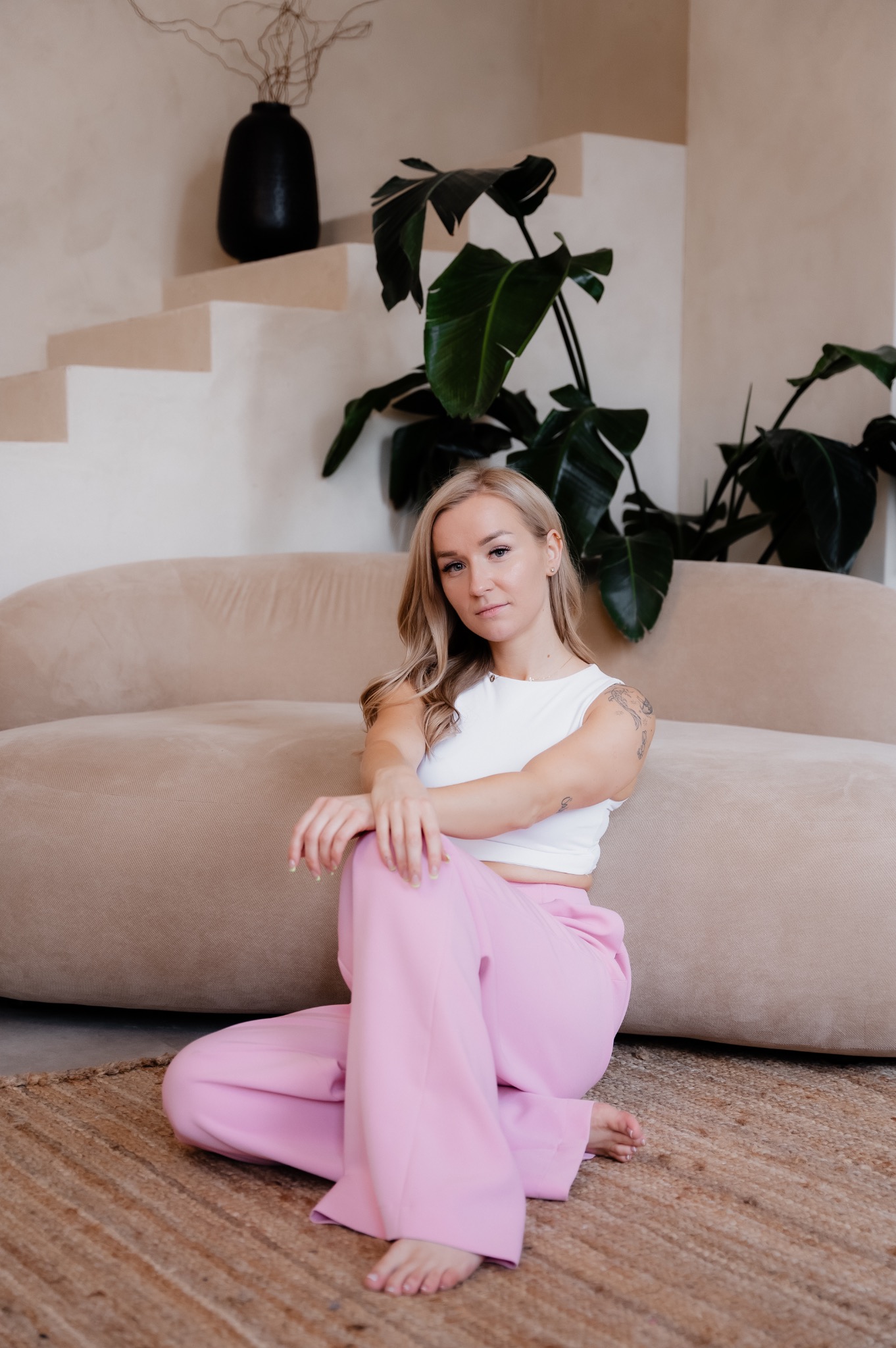 Kobieta siedzi na podłodze w różowych spodniach i białym topie, opierając się o beżową sofę w minimalistycznym wnętrzu z roślinami i dekoracyjnymi schodami.