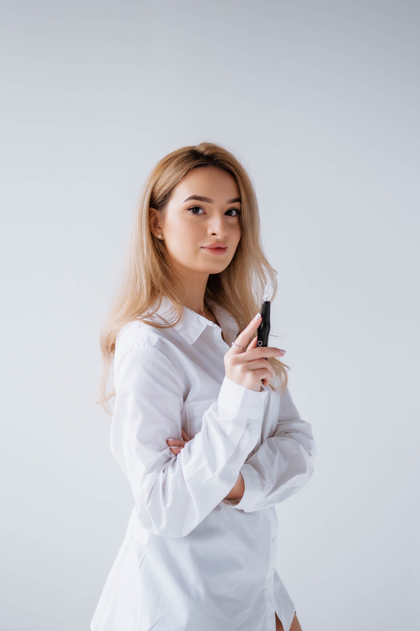 Blondynka w białej koszuli, z rękami skrzyżowanymi na piersiach, trzyma czarny aplikator kosmetyczny z przezroczystą końcówką na tle jasnego studia.
