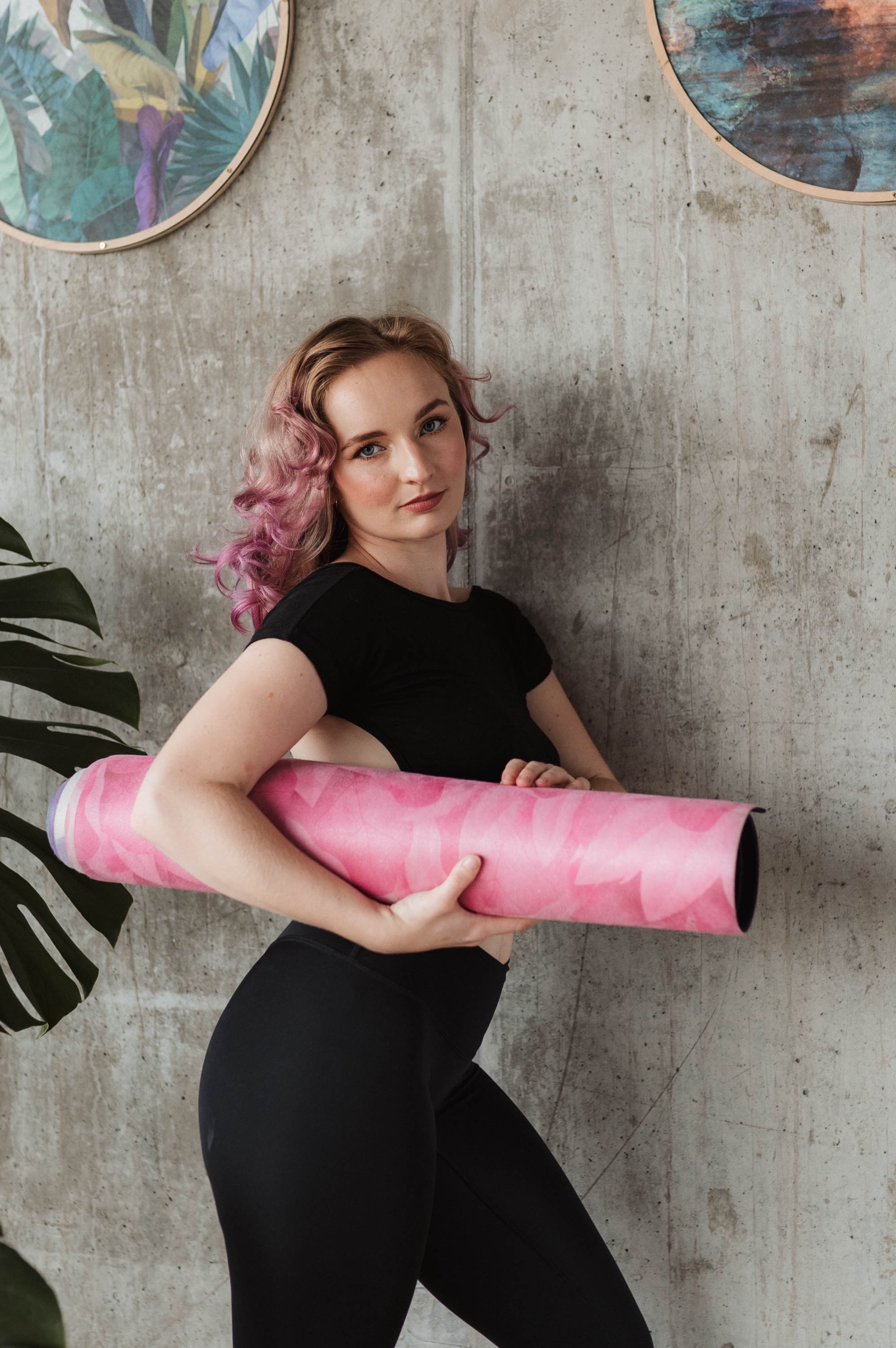 Kobieta z różowymi końcówkami włosów, ubrana w czarny strój sportowy, trzyma zwiniętą różową matę do ćwiczeń na tle betonowej ściany z dekoracyjnymi obrazkami i rośliną.