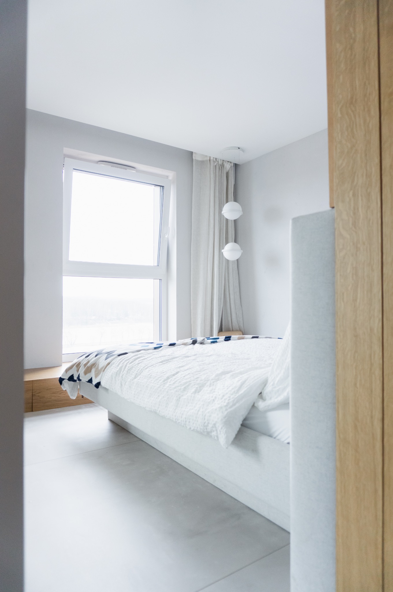 Jasna sypialnia z dużym oknem, białymi ścianami i podłogą, minimalistycznym łóżkiem z białą pościelą i wzorzystą narzutą, oraz wiszącymi kulistymi lampami.