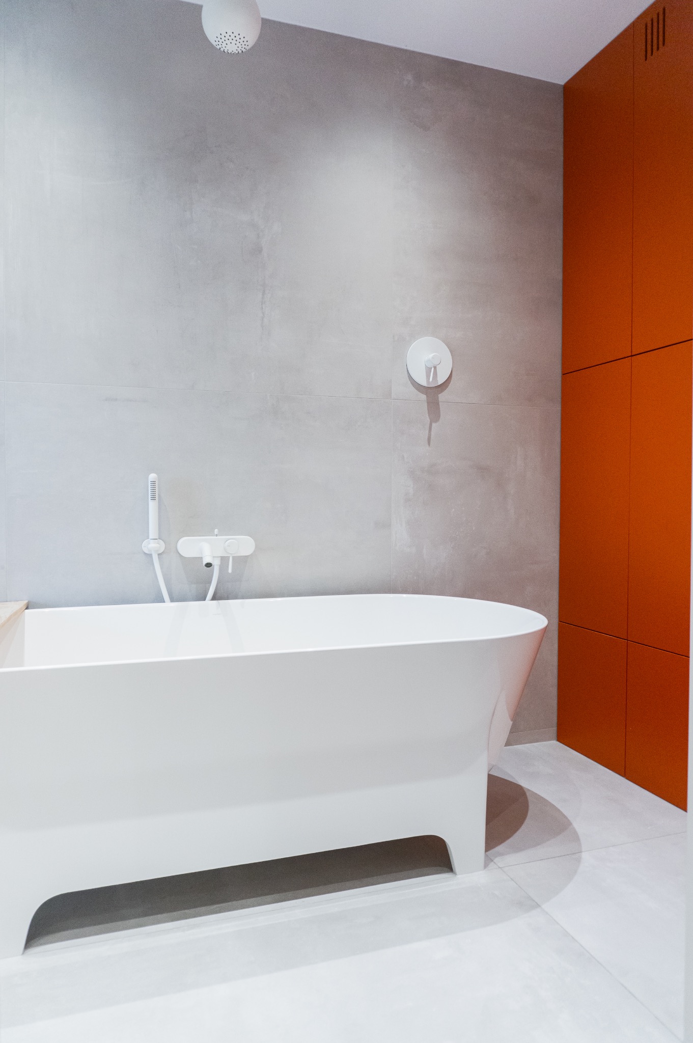 Minimalistyczna łazienka z wolnostojącą białą wanną, szarą ścianą z armaturą i pomarańczową szafką.