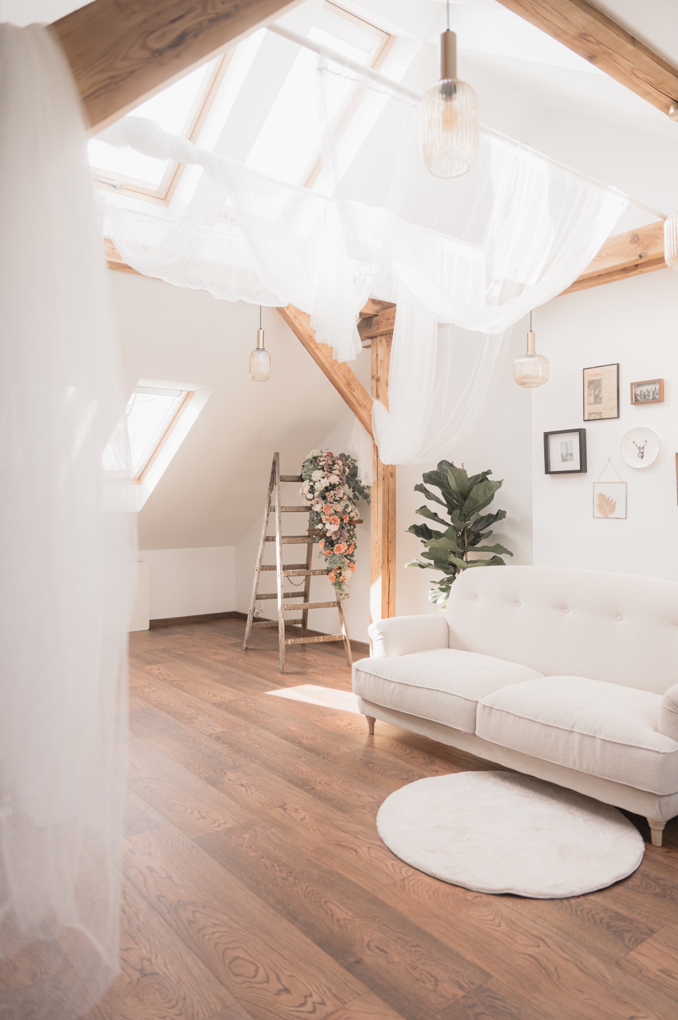 Jasne, przestronne poddasze z drewnianą podłogą, sofą w stylu vintage, drabiną z kwiatami i dekoracyjnymi lampami wiszącymi pod oknami dachowymi.