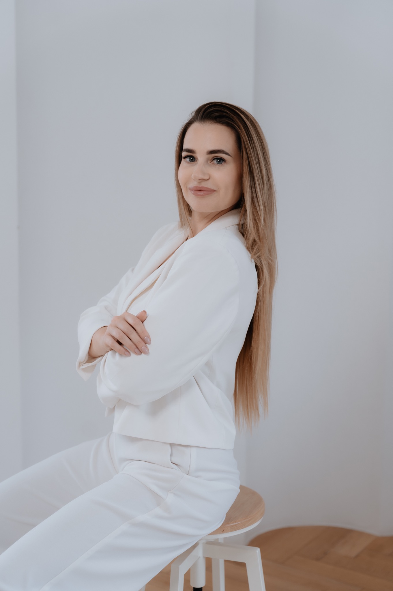 Kobieta w białym garniturze siedząca na drewnianym stołku, z rękami skrzyżowanymi na piersiach i uśmiechem, na jasnym tle.