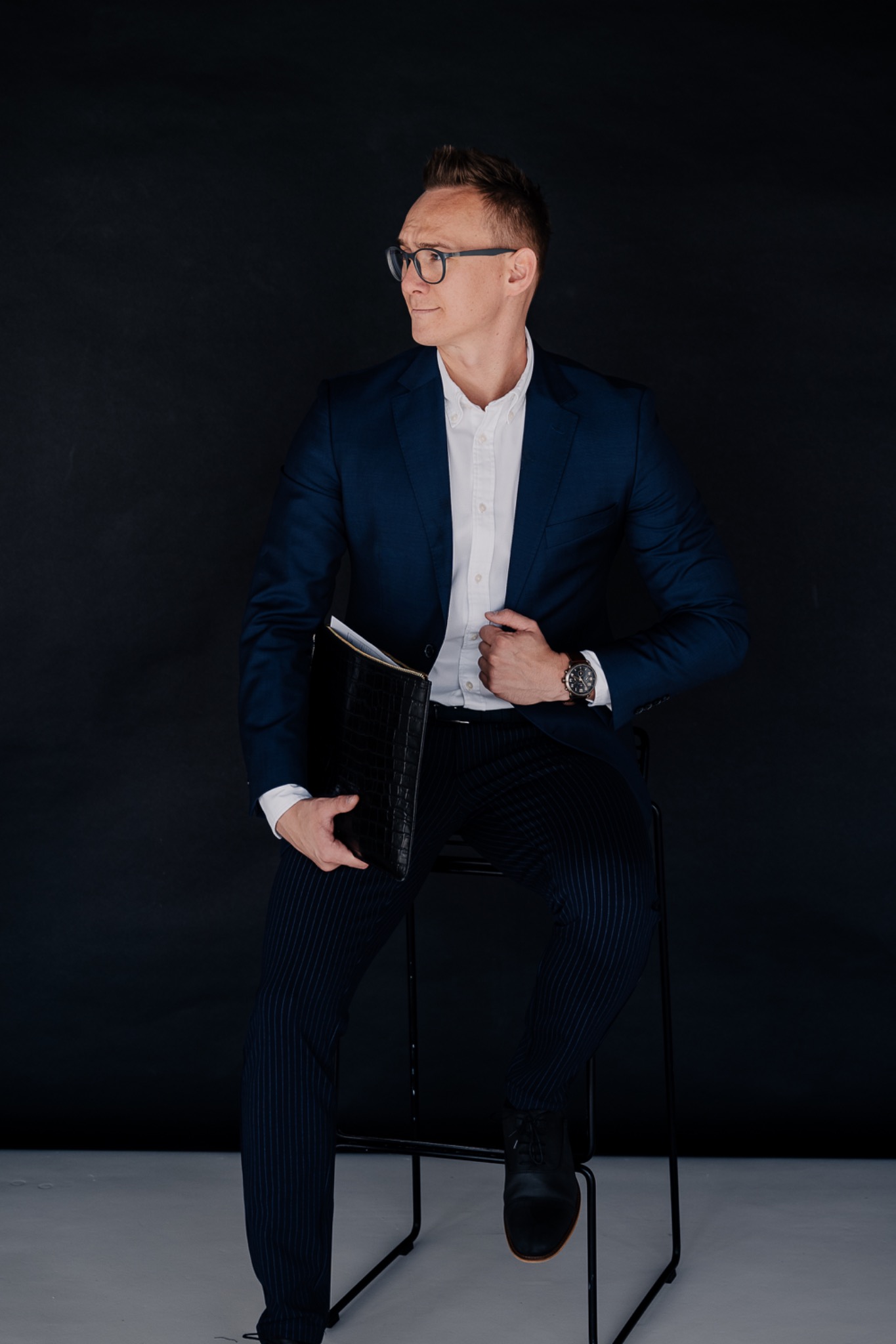 Mężczyzna w okularach, granatowym garniturze i białej koszuli, siedzący na krześle z czarną aktówką, spoglądający w bok.