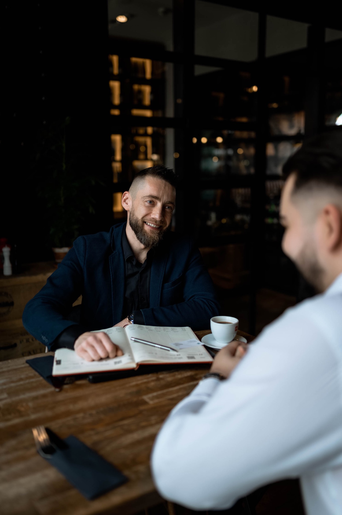 Mężczyzna w ciemnej marynarce uśmiecha się podczas spotkania biznesowego w kawiarni, notując coś w otwartym kalendarzu obok filiżanki kawy i długopisu.