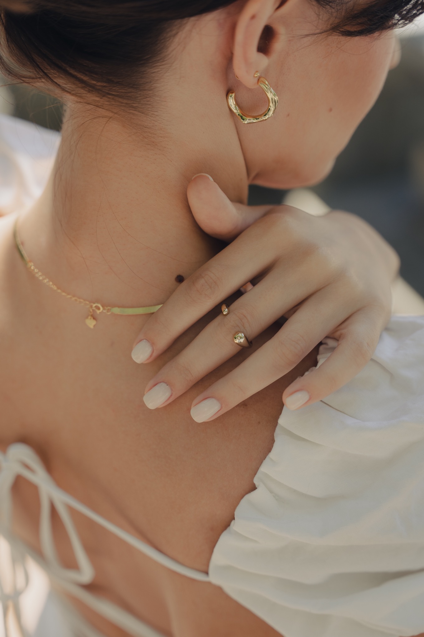Kobieta w białej bluzce prezentuje złotą biżuterię: kolczyki, naszyjnik i pierścionek z kamieniem, dłoń delikatnie dotyka szyi.