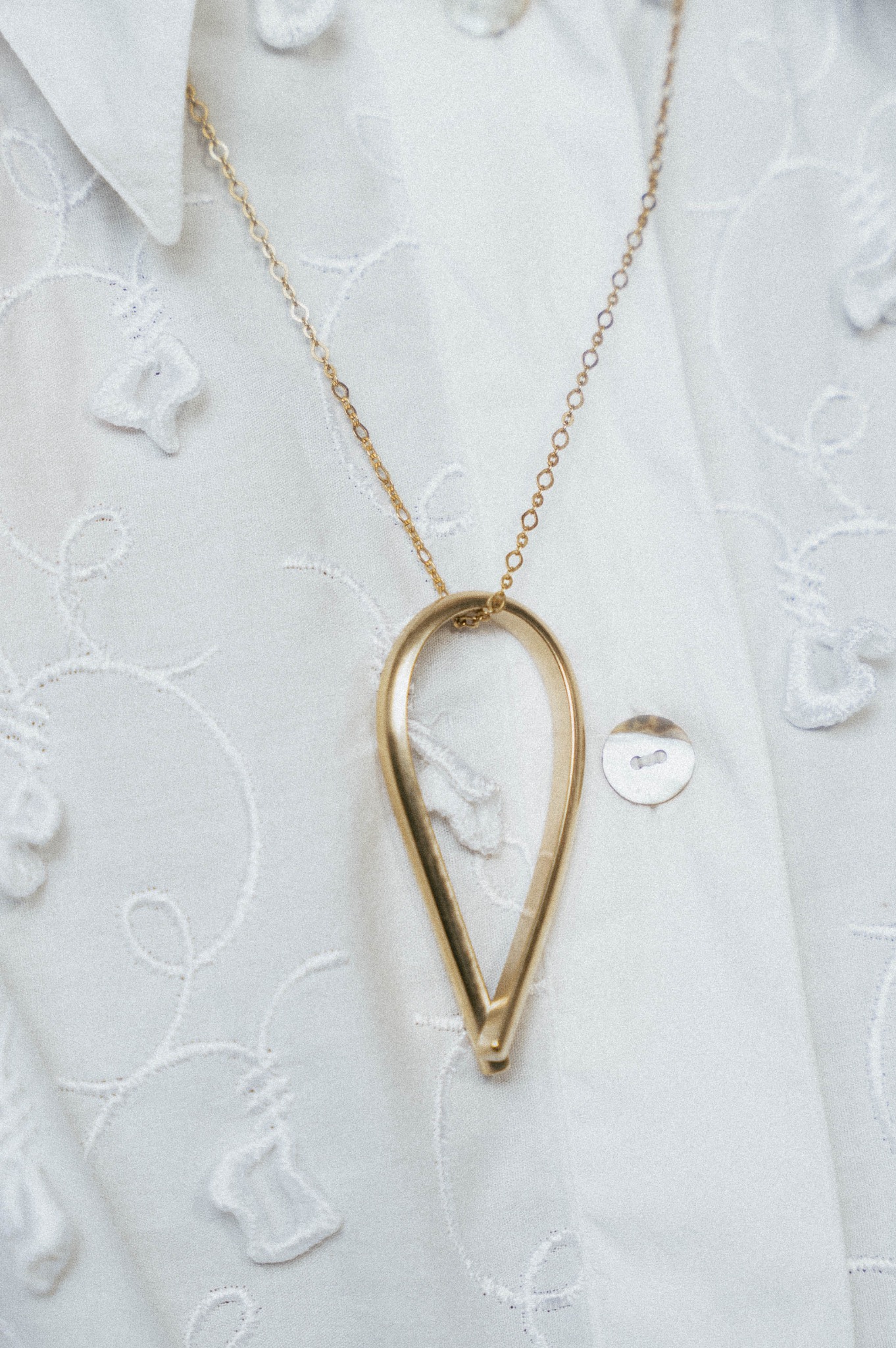 Złoty naszyjnik z geometrycznym wisiorkiem w kształcie łzy, leżący na białej koszuli z haftowanymi kwiatami i perłowymi guzikami.