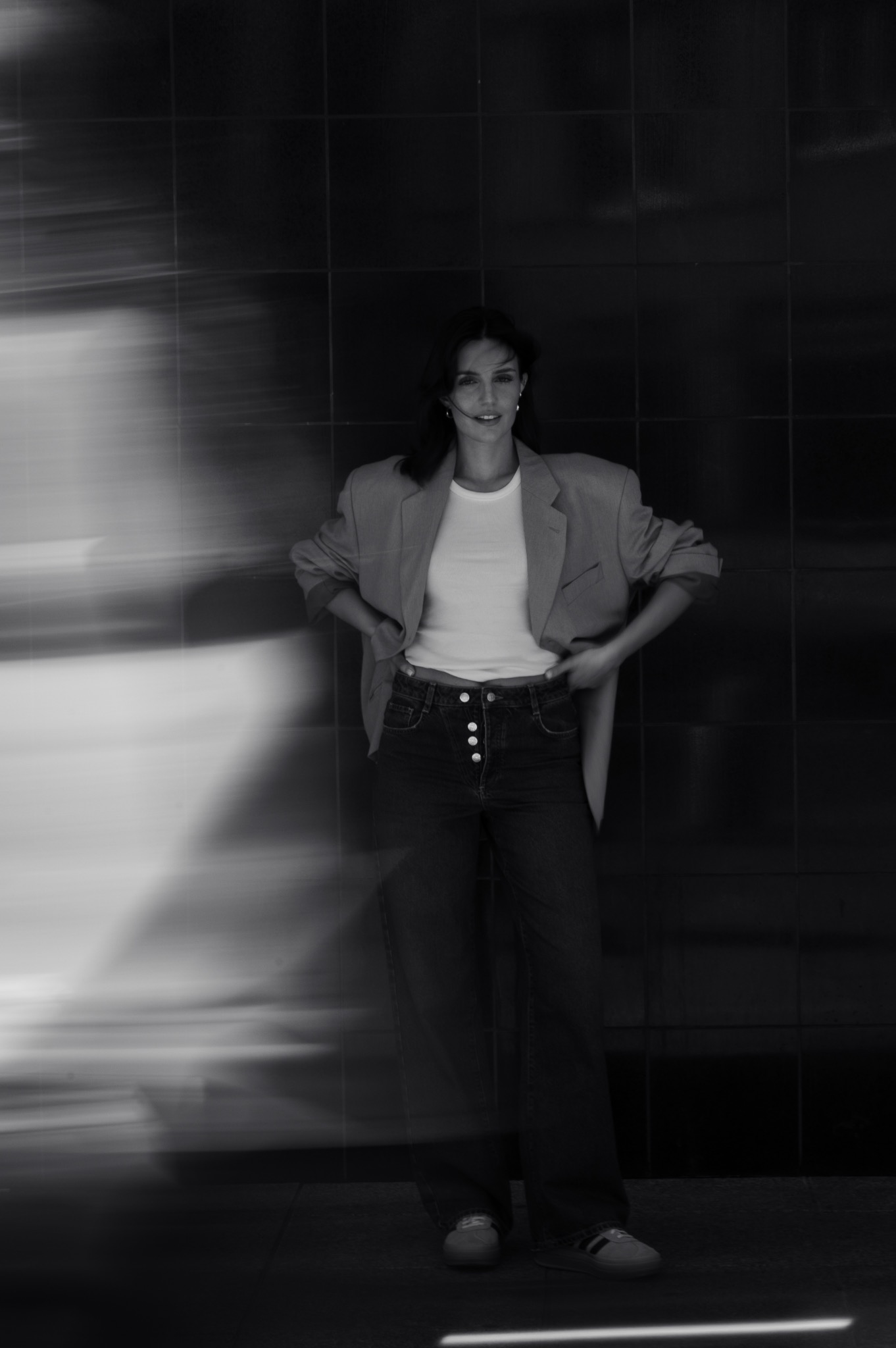 Portret kobiety w luźnym blazerze i jeansach z guzikami, pozującej na tle ciemnej, kafelkowanej ściany z efektem rozmycia po lewej stronie.