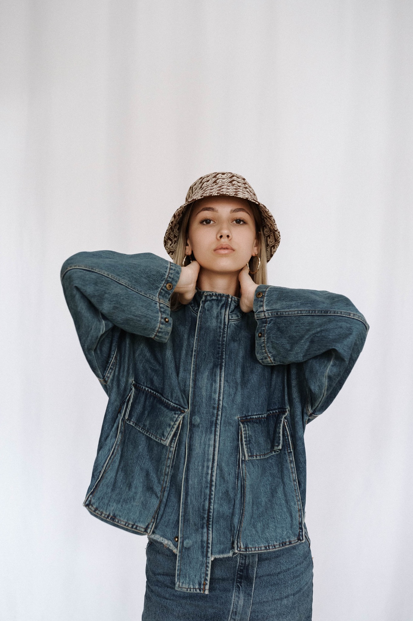 Młoda kobieta w dżinsowej kurtce i wzorzystym kapeluszu typu bucket, pozuje z rękami na karku, patrząc prosto w obiektyw na jasnym tle.