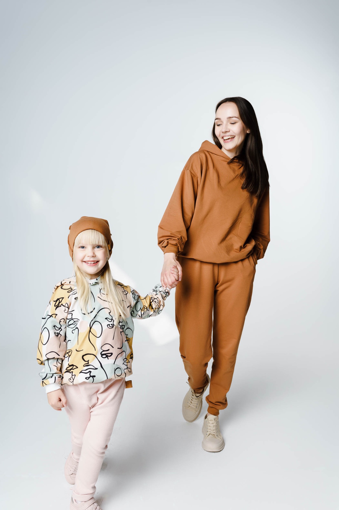 Uśmiechnięta kobieta i dziewczynka w pasujących dresach, trzymające się za ręce na białym tle. Dziewczynka ma na sobie czapkę i bluzę z abstrakcyjnym wzorem twarzy.