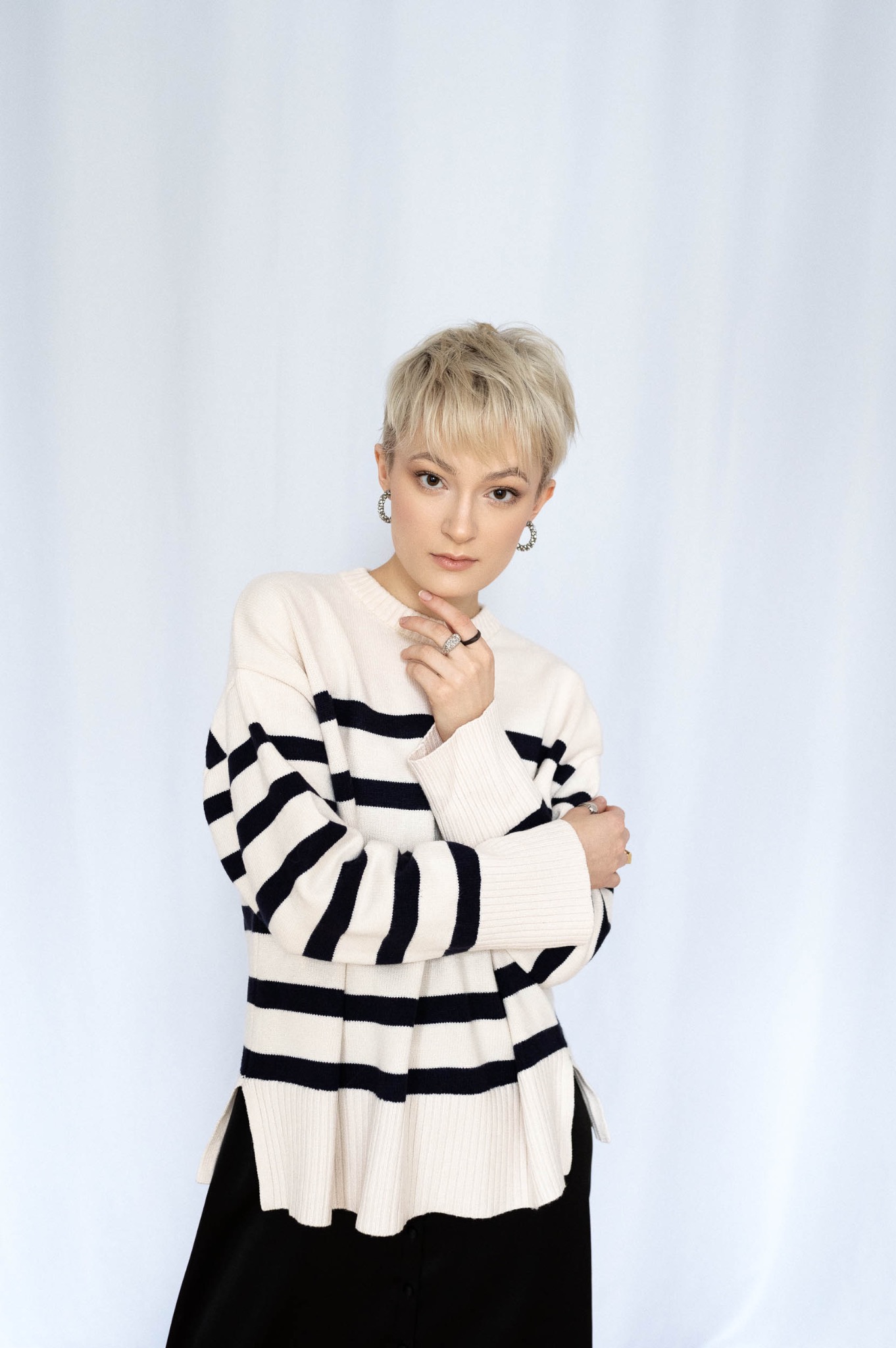 Portret kobiety z krótkimi blond włosami w swetrze w czarno-białe paski, z ręką pod brodą, na białym tle.