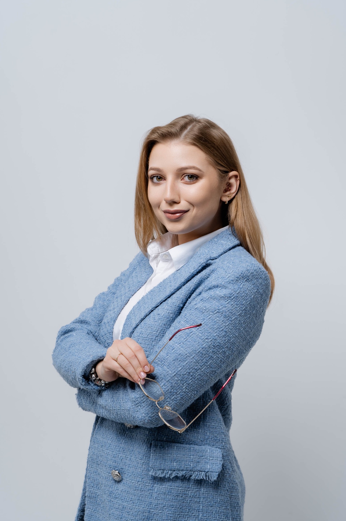 Uśmiechnięta blondynka w błękitnym tweedowym żakiecie i białej koszuli, trzymająca okulary w ręce, pozuje na jasnoszarym tle.