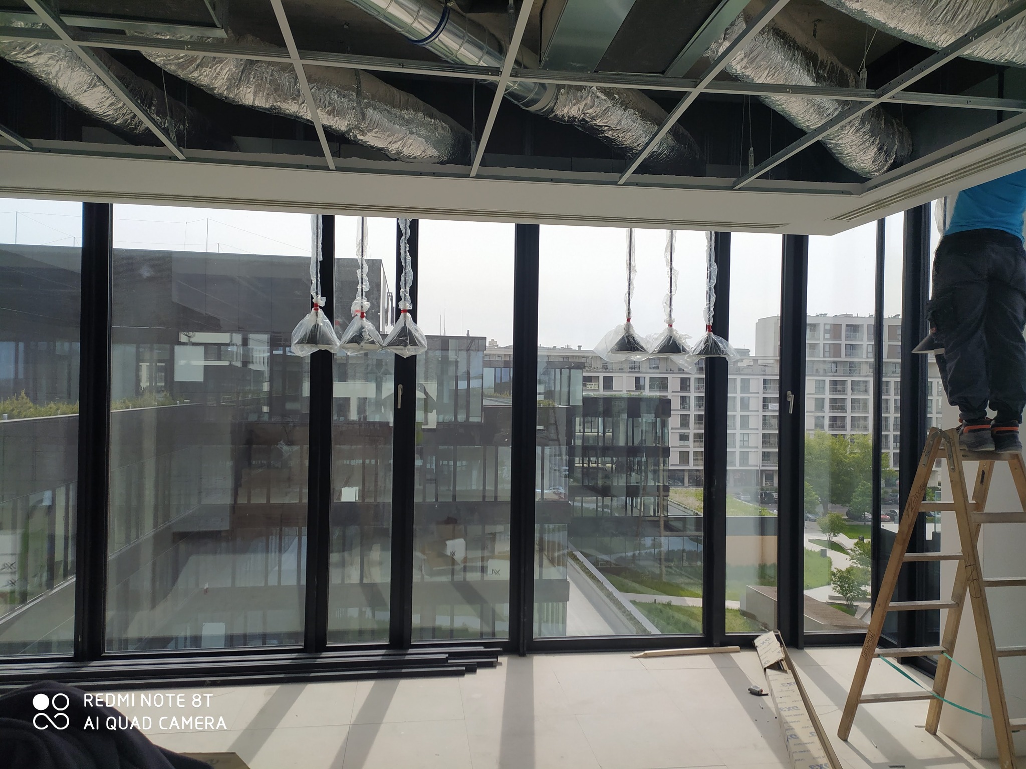 Pracownik na drabinie, montaż sufitu podwieszanego z widocznymi elementami instalacji wentylacyjnej i wiszącymi lampami w folii ochronnej, na tle okien z widokiem na budynki miejskie.