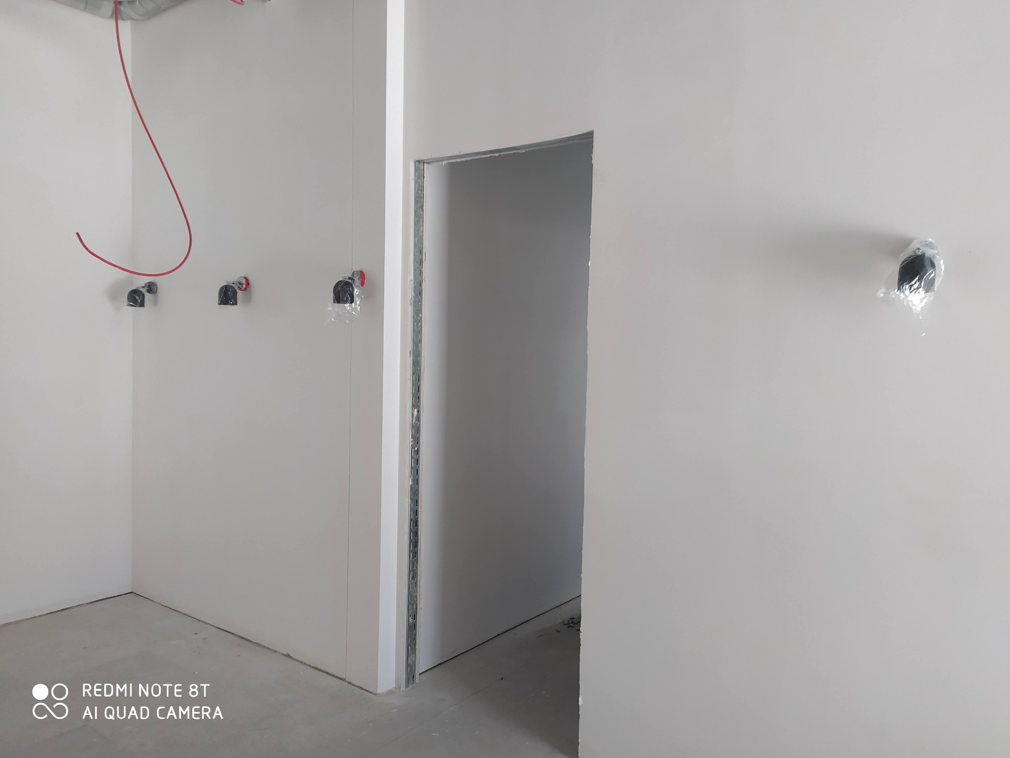 Surowy stan wnętrza w trakcie prac wykończeniowych: widoczne wystające przewody elektryczne i puszki zabezpieczone folią, szare ściany i surowy otwór drzwiowy.
