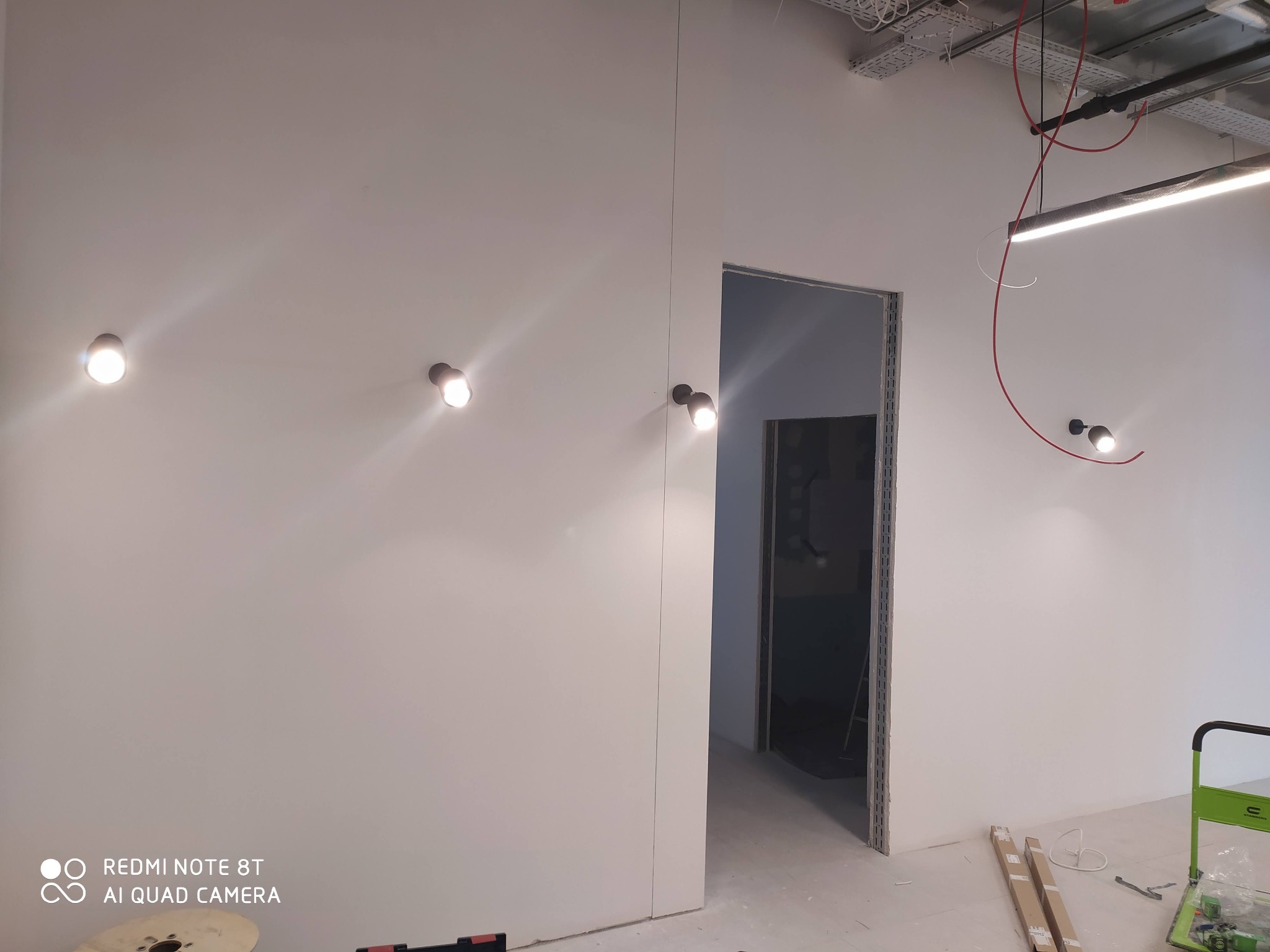 Wnętrze w trakcie wykańczania z zamontowanym punktowym oświetleniem ściennym, widoczne kable pod sufitem i surowy stan wykończenia ścian oraz podłogi.