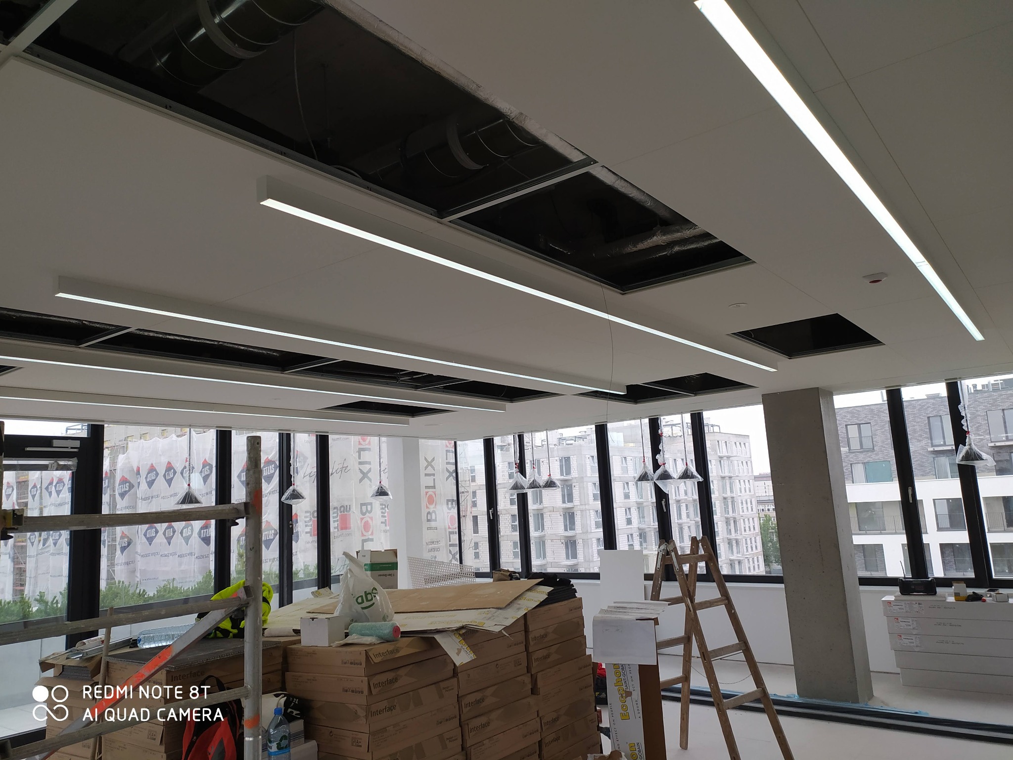 Wnętrze biurowe w trakcie wykończenia, widoczne liniowe lampy LED zamontowane w suficie podwieszanym, część paneli sufitowych jeszcze nie zamontowana, rusztowanie, drabina i materiały budowlane...