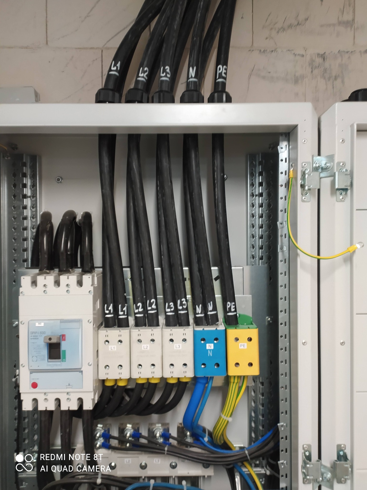 Otwarta szafa elektryczna z zainstalowanymi zabezpieczeniami, czarnymi przewodami oznaczonymi L1, L2, L3, N, PE, oraz kolorowymi złączkami.