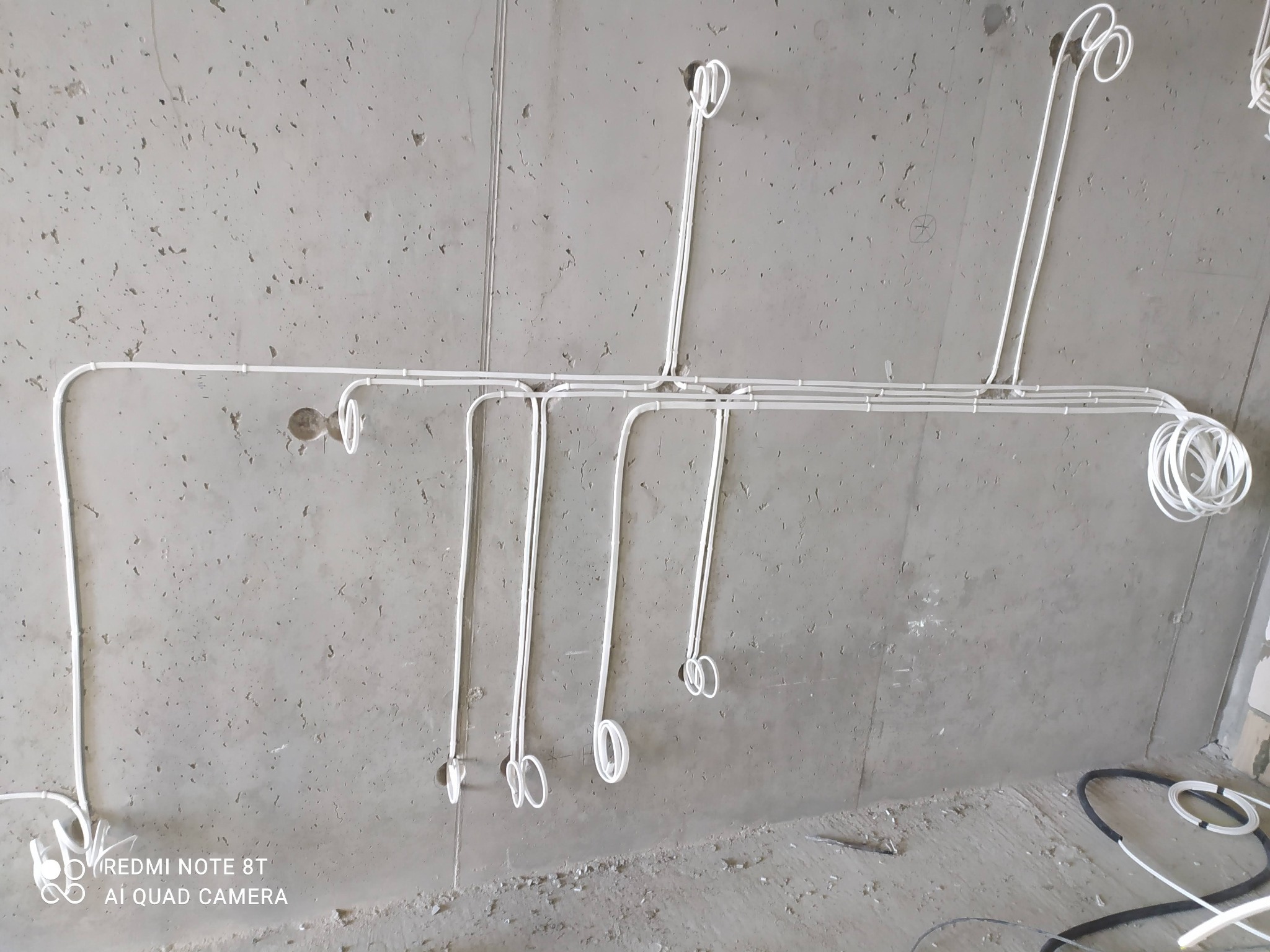 Białe przewody elektryczne zamocowane do surowego, betonowego sufitu i ściany, przygotowane do podłączenia oświetlenia, z pętlami zwisającymi w miejscach przyszłych punktów świetlnych.