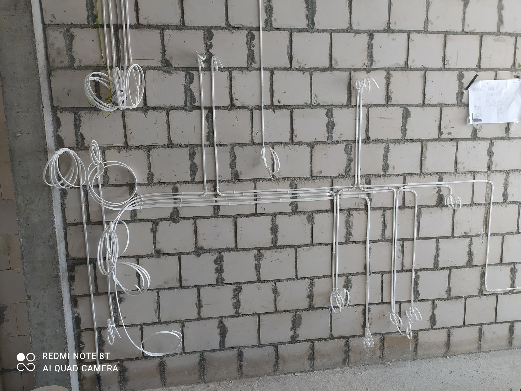 Białe kable elektryczne prowadzone po surowej ścianie z bloczków betonowych, z widocznym planem na kartce przyklejonej do ściany.